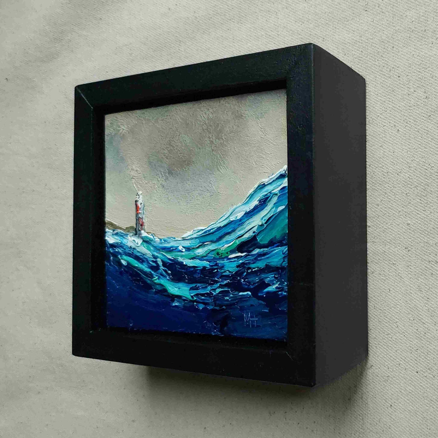 Framed painting of a lighthouse surrounded by waves on a textured wall viewed from the side.