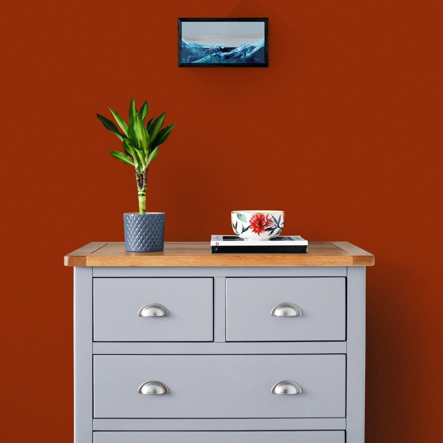 Wave painting above grey dresser with wooden top against a red wall with a plant and decorative items.